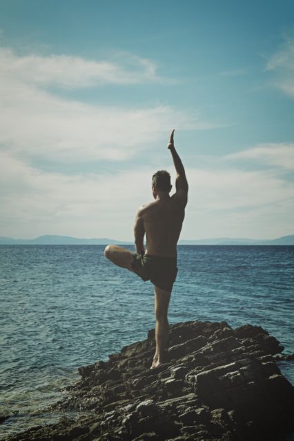 Man practicing yoga on a rocky beach by the ocean, showcasing balance and tranquility. Ideal for use in wellness and meditation-related promotions. Perfect for articles or advertisements about outdoor fitness, healthy living, and the benefits of yoga. Also suitable for travel magazines focusing on beautiful and serene beach destinations.