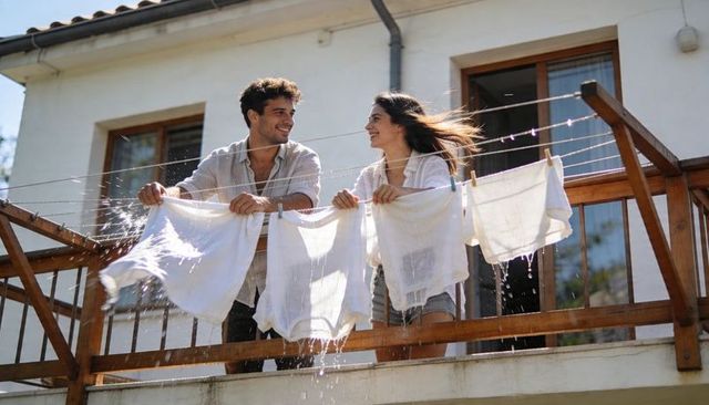 Young couple hanging wet linens on sunny balcony laughing and shaking towels