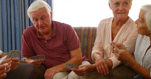 Seniors sitting on a couch and engaging in a card game, exhibiting enjoyment of social interaction. Ideal for use in articles about leisure activities for the elderly, advertisements for retirement communities, senior lifestyle pieces, and content promoting social engagement among older adults.