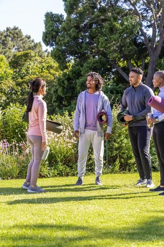 Friends chatting outdoors holding yoga mats in sunlit park for group fitness and wellness