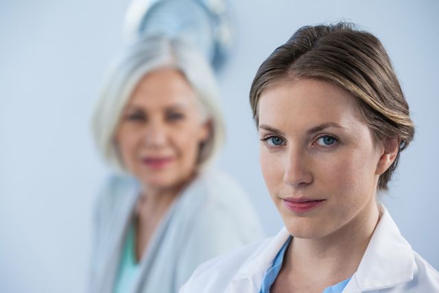 Young female doctor smiling confidently in a medical office, with an older patient in the background. Ideal for use in healthcare advertisements, medical websites, and promotional materials for clinics and hospitals. Highlights professionalism and trust in healthcare settings.