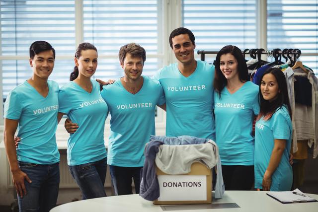Group of diverse volunteers standing together in a donation center, smiling and wearing blue shirts with 'Volunteer' written on them. They are gathered around a donation box filled with clothes. Ideal for use in materials promoting community service, charity events, nonprofit organizations, and volunteer recruitment.