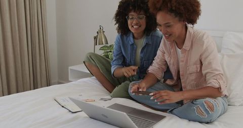 Mid adult women collaborating on bed with laptop and smartphone, cozy home teamwork