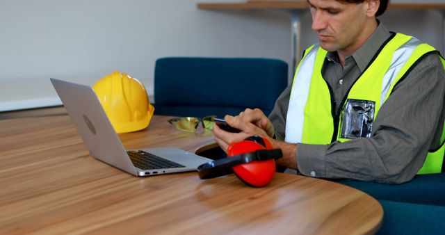 Construction worker reviews plans on a laptop at the office. Safety gear like a helmet and ear protection are placed on the table, indicating a focus on workplace safety.