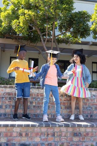 Joyful diverse graduation trio wearing mortarboards holding diplomas on brick wall