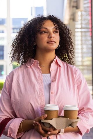 Confident african woman holding two takeaway coffees in carrier by modern glass building