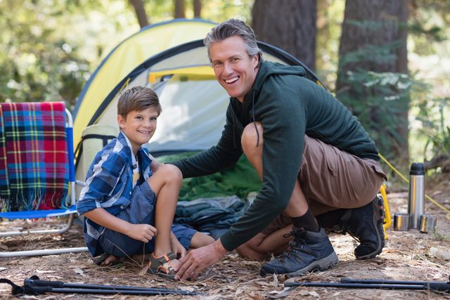 Father and son enjoying a camping trip in the forest. They are kneeling by a tent, smiling at the camera. This image can be used for promoting outdoor activities, family bonding, travel destinations, and camping gear.