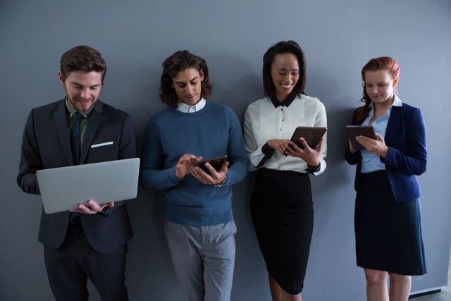 Group of business professionals standing against a gray wall, using various electronic devices such as laptops and tablets. Ideal for illustrating modern workplace technology, teamwork, and corporate communication. Suitable for business presentations, websites, and marketing materials focusing on digital collaboration and productivity.