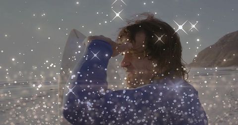 Windblown surfer shading eyes holding white surfboard on wet sand with sparkle bokeh