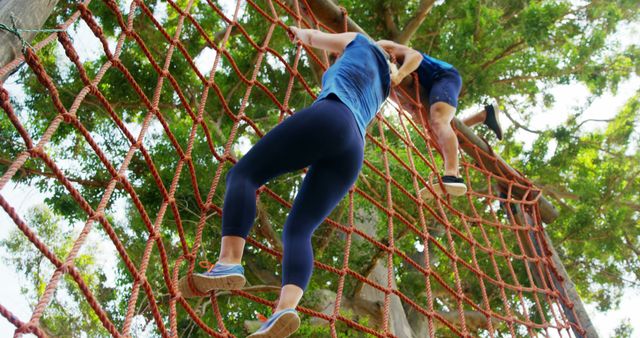 A young Caucasian woman is actively climbing a rope net at an outdoor obstacle course, with copy space. Her athletic attire and focused demeanor suggest she's engaged in a fitness challenge or adventure activity.