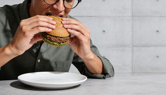 Man biting sesame burger wearing glasses and olive shirt at concrete table