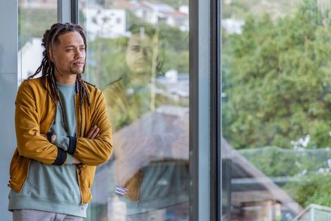 African american man standing by window, gazing out with reflection, mustard jacket
