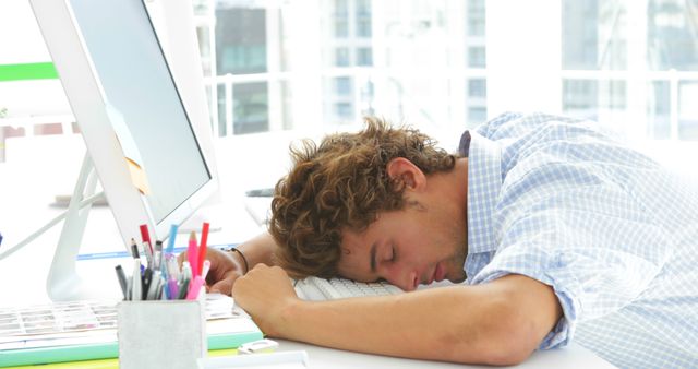 Young professional man sleeping with his head on keyboard at his desk. Concept of overwork, stress, and employee burnout. Useful for materials on workplace stress, mental health, and productivity challenges.