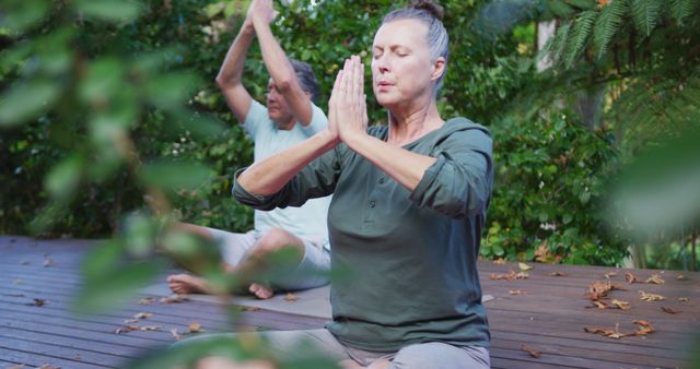 Senior couple engaged in yoga and meditation routine on wooden deck surrounded by greenery. Ideal for advertisements and promotions related to healthy lifestyles, senior fitness programs, outdoor activities, and wellness retreats.