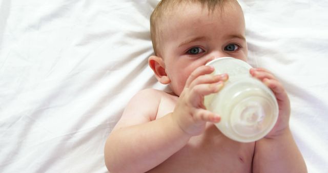 This image captures a baby comfortably lying on a bed, drinking from a bottle. The baby appears calm and content while feeding. It can be used for parenting blogs, baby care products, advertisements for infant formula, or articles about child nutrition.