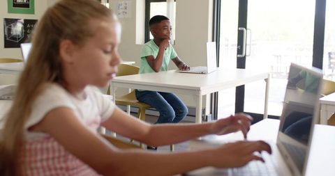 Diverse elementary students typing and thinking on laptops in sunlit classroom