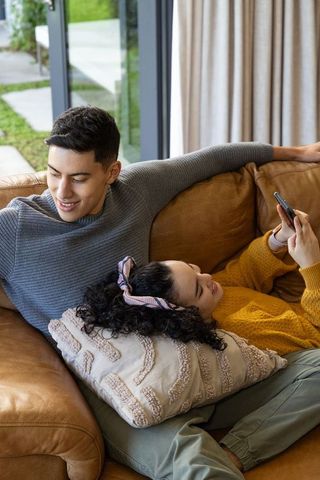 Young couple relaxing on brown leather sofa browsing smartphone in bright living room