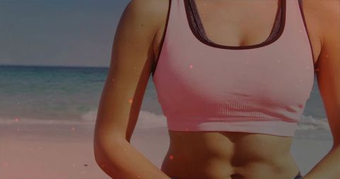 Fit woman standing on beach wearing pink sports crop top revealing toned abs