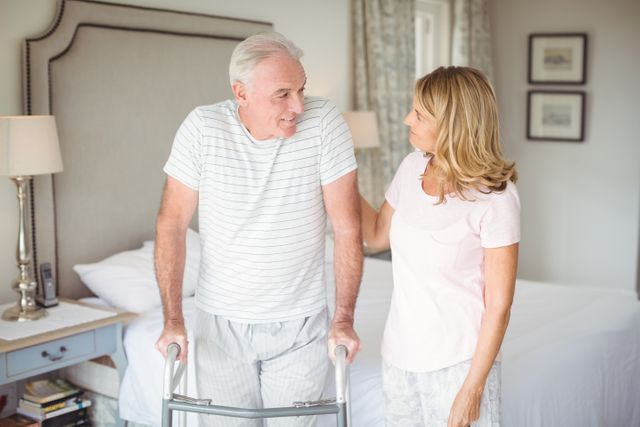 Senior woman assisting elderly man using a walker in a bedroom. Ideal for use in healthcare, caregiving, senior living, and family support contexts. Highlights themes of aging, recovery, and family care.