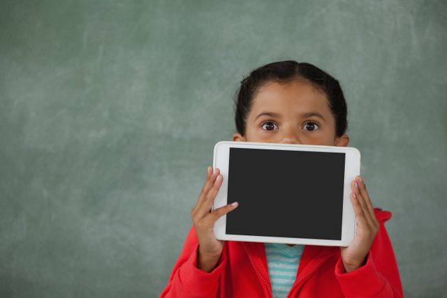 Young girl holding digital tablet in front of her face against chalk board