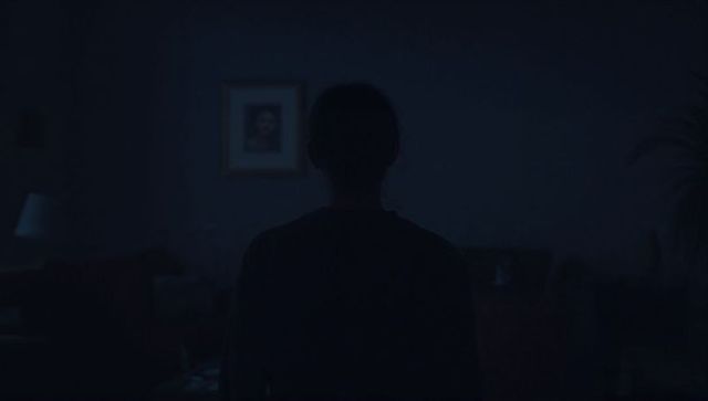 Solitary silhouette standing in dim living room facing framed portrait, moody interior