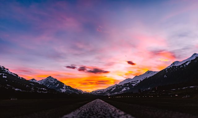 Road illuminated by vivid sunset colors, lined with snow-capped mountains. Perfect for travel, outdoor adventure, and scenic views content.