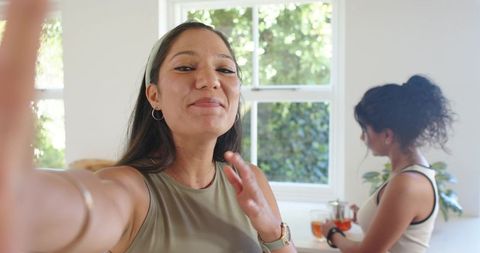 Indian woman taking selfie in bright kitchen while friend pouring tea at counter
