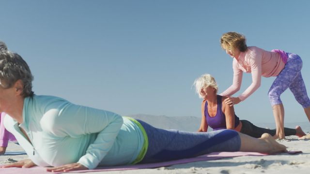 A group of senior women engaging in a yoga session on a serene beach with the blue sky and sea in the background. One of the women is serving as an instructor, assisting the others with poses. This scene epitomizes relaxation, wellness, and a healthy lifestyle. It works well for promoting fitness activities, wellness programs, or advertisements emphasizing outdoor, carefree living for seniors.