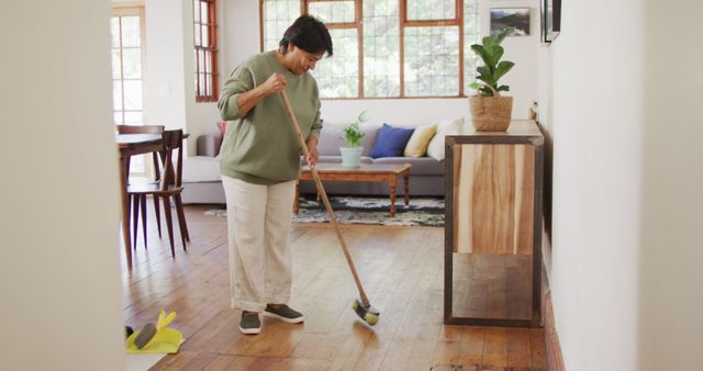 Image of a senior woman sweeping a hardwood floor in a brightly lit living room. This photo can be used to illustrate concepts related to domestic life, senior activities, home cleaning routines, and maintaining a tidy living space. Suitable for articles on home organization, cleaning tips, and elder lifestyle.
