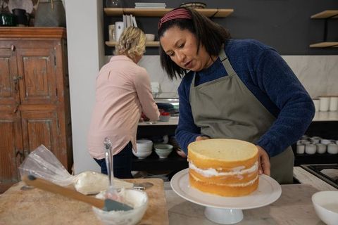 Home bakers assembling layered sponge cake on white stand in rustic kitchen with frosting tools