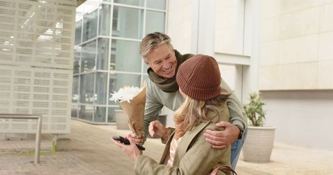 Mature man surprising partner with bouquet while embracing on modern urban plaza