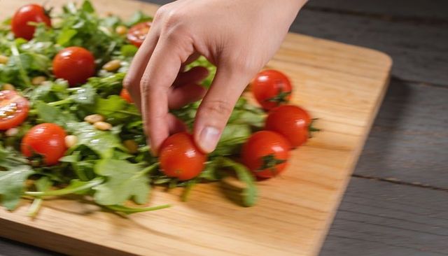Hand picking cherry tomato on wooden cutting board with arugula and pine nuts