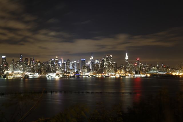 Dramatic night view of New York City skyline with buildings reflecting on water. Perfect for travel promotions, cityscape backgrounds, urban lifestyle content, tourism advertising.
