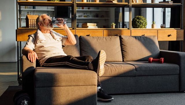 Sunlit senior drinking water on gray sofa after home workout | active aging hydration