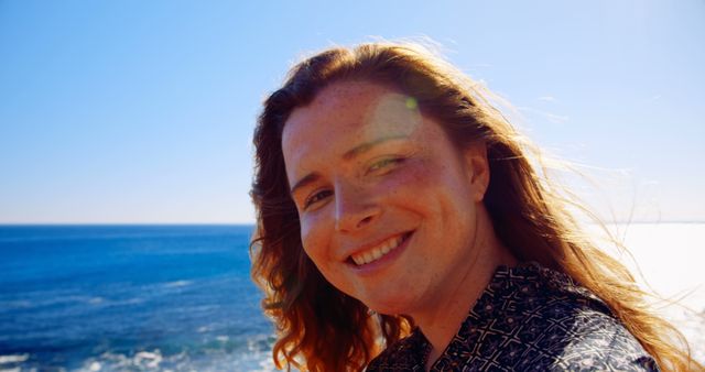 A woman with light brown hair is smiling brightly near an ocean with a clear blue sky in the background. This evokes a sense of happiness, joy, and tranquility. Ideal for use in travel blogs, promotional materials for seaside destinations, wellness retreats, or to depict serene outdoor life on a sunny day.