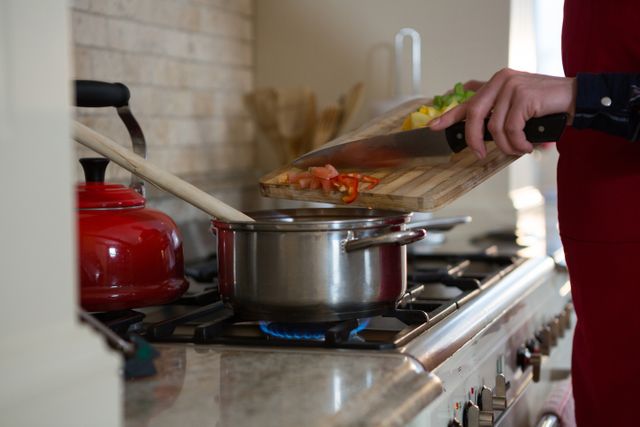 Mid section of woman cooking vegetables in a modern kitchen at home. She is adding chopped vegetables from a wooden chopping board into a pot on the stove. A red kettle is visible in the background. Ideal for use in articles or advertisements related to home cooking, healthy eating, kitchen appliances, and lifestyle blogs.