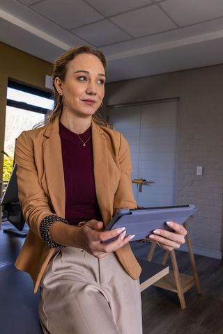 Young professional woman sitting on desk edge holding tablet and looking thoughtfully