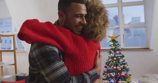 A happy couple embracing in a cozy apartment with festive Christmas tree glowing in the background. Ideal for holiday greeting cards, festive season promotions, advertisements for home decor, or relationship blog posts.