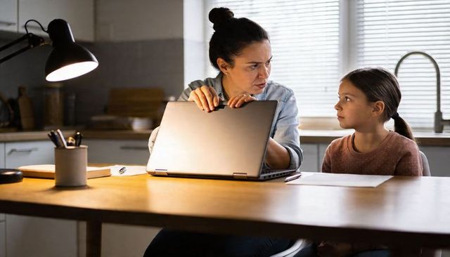 Parent adjusting laptop while helping child with homework at kitchen table during evening