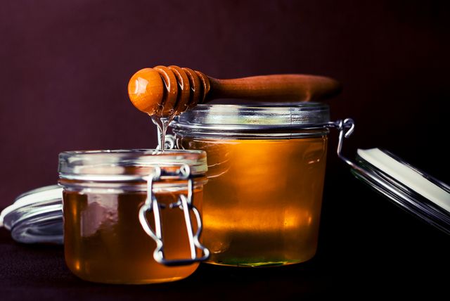 Glowing honey jars with wooden drizzler on dark background. Ideal for use in culinary blogs, health food packaging design, kitchen decor, nutritional supplement advertisements, and recipe websites.