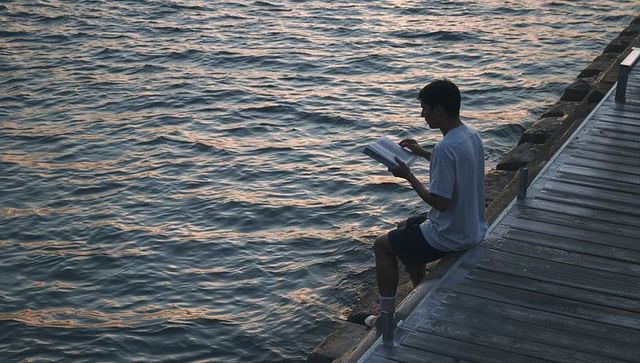 Young man reading on pier at sunset, contemplative solitude by calm water with golden hour light