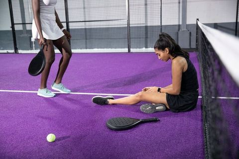 Female padel player sitting on purple court checking injured leg with teammate holding paddle