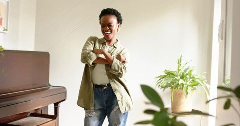 Joyful woman smiling with arms crossed in sunlit living room near piano and plants