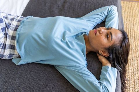 Asian woman lying relaxed on daybed wearing light blue top and plaid pajama pants, cozy minimalist b
