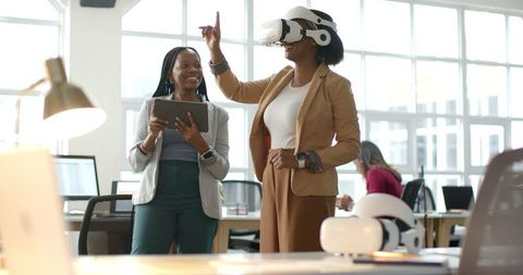 Two African American businesswomen using VR headset in modern office collaborating with tablet