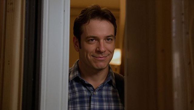 Smiling man in doorway wearing plaid shirt and backpack welcoming into cozy hallway