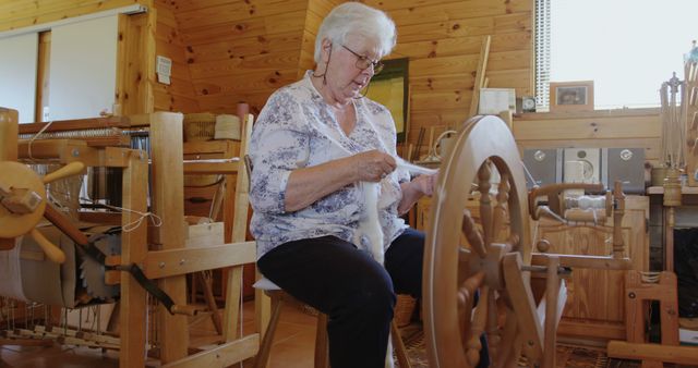 Senior Caucasian woman spins yarn on an antique wheel at home. Her skilled hands demonstrate a traditional craft in a cozy, rustic setting.