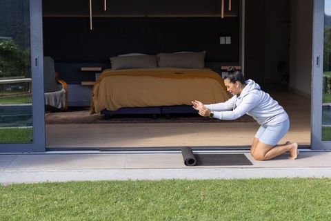 Woman kneeling prepping yoga mat at open bedroom threshold on modern terrace