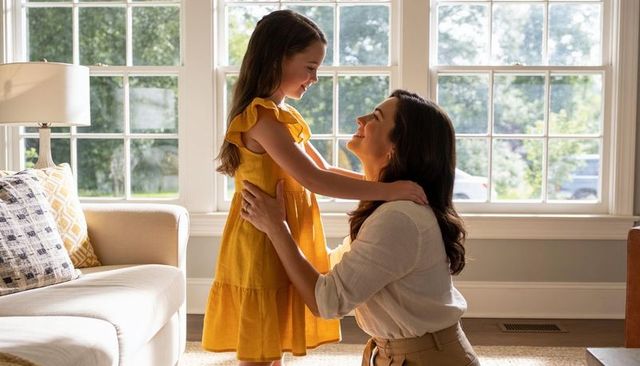 Mother kneeling embracing daughter in sunlit living room, warm family connection