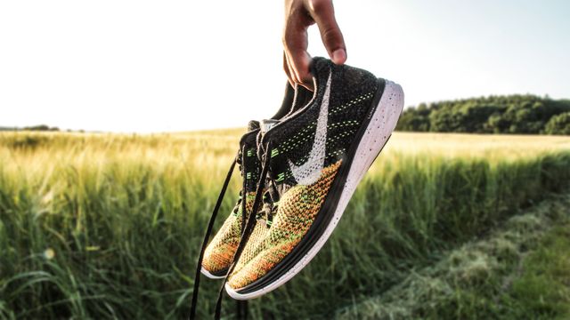 Person holding pair of multicolored running shoes against backdrop of open field during sunset. Perfect for fitness, outdoor activities, running, healthy lifestyle, and exercise-related themes. Suitable for promoting sportswear, fitness equipment, nature photos, or countryside settings.